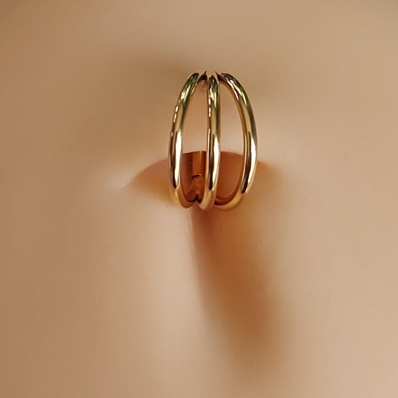 May include: Gold triple ring belly button piercing. The ring is made of three interlocking circles.