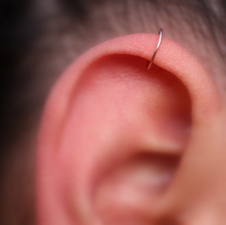 May include: Close-up of a person's ear with a small, silver hoop earring in the upper cartilage.
