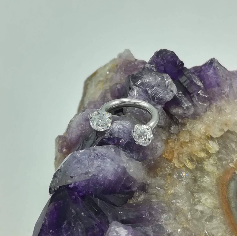 May include: A silver horseshoe-shaped ring with two clear round gemstones on either end. The ring is resting on a purple and white crystal.
