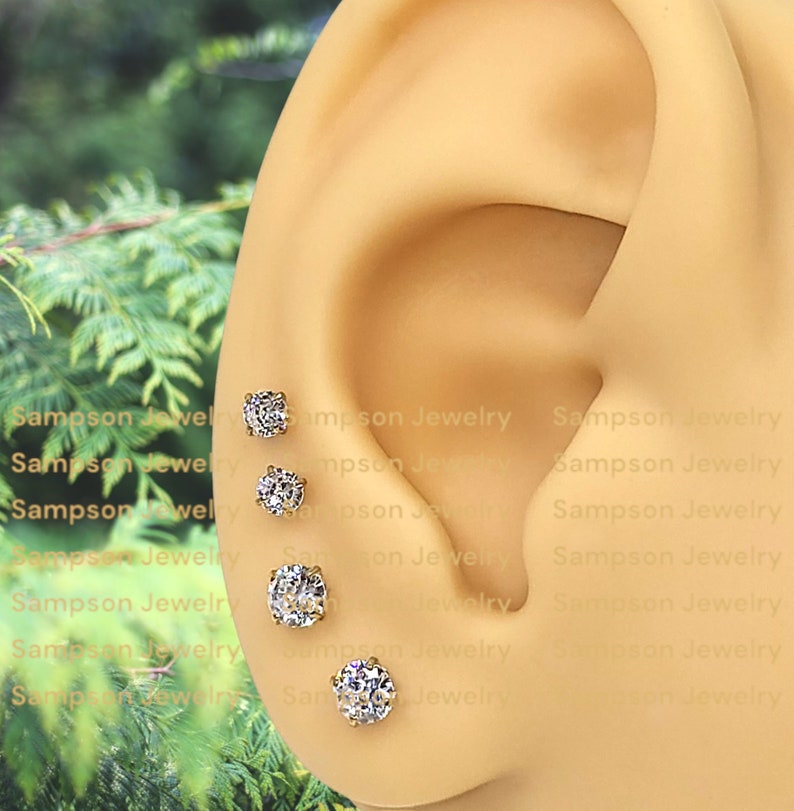 May include: Four gold stud earrings with round, clear cubic zirconia stones. The earrings are arranged in a row on a model's ear.