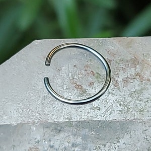 May include: A silver nose ring hoop, a type of body jewelry, is shown on a white background.