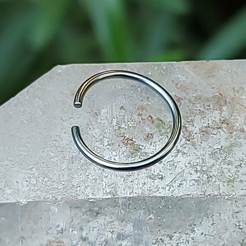 May include: A silver nose ring hoop, a type of body jewelry, is shown on a white background.