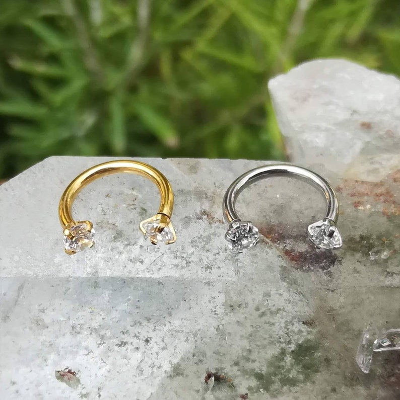May include: Two circular barbell earrings with clear gemstones. One earring is gold and the other is silver.