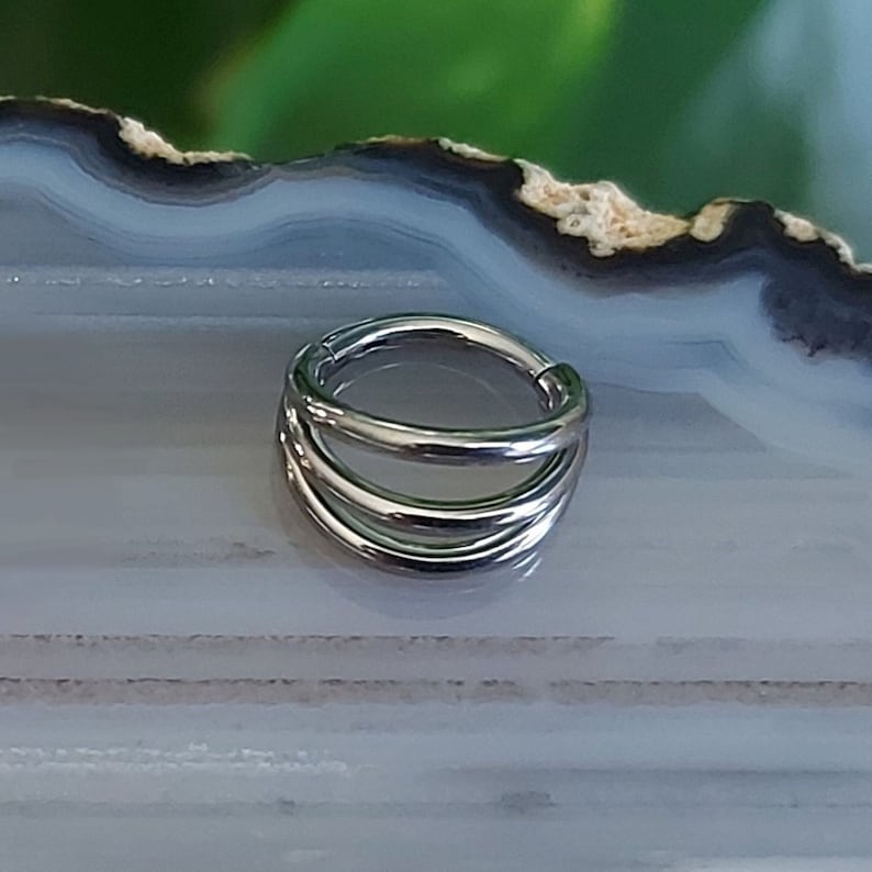 May include: Silver triple hoop septum ring. The ring is made of three hoops that are stacked on top of each other. The ring is a popular choice for septum piercings.