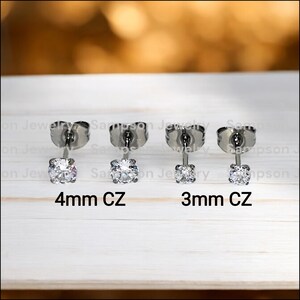 May include: Two pairs of silver stud earrings with clear cubic zirconia stones. The left pair has 4mm stones and the right pair has 3mm stones. The earrings are on a white wooden surface.