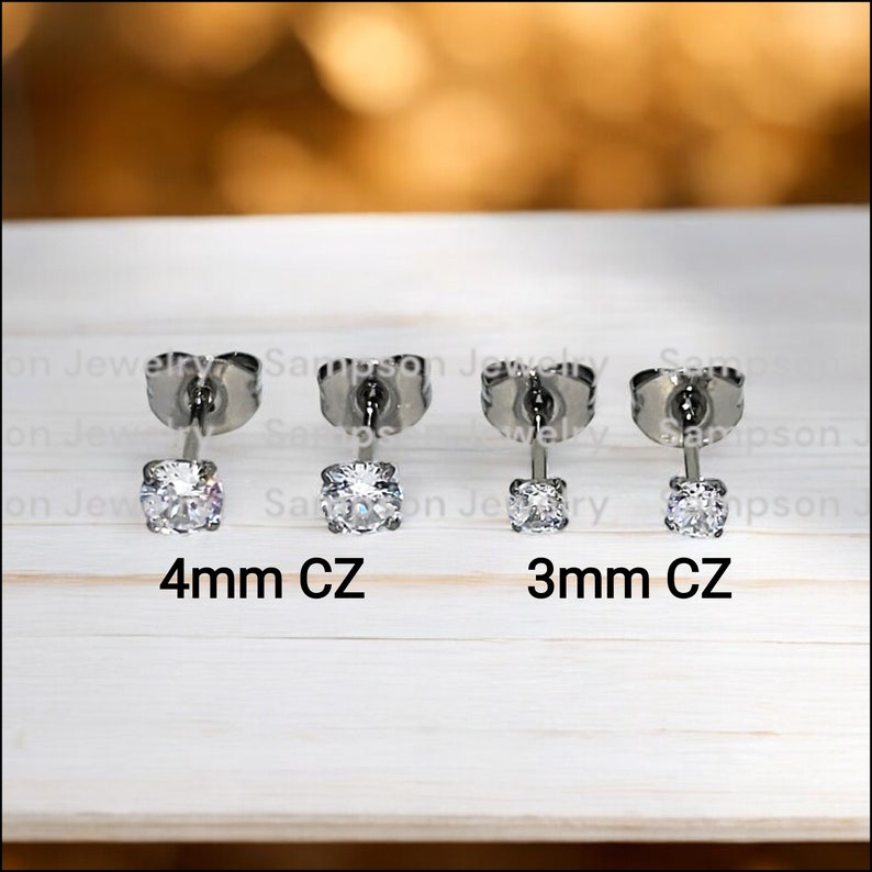 May include: Two pairs of silver stud earrings with clear cubic zirconia stones. The left pair has 4mm stones and the right pair has 3mm stones. The earrings are on a white wooden surface.