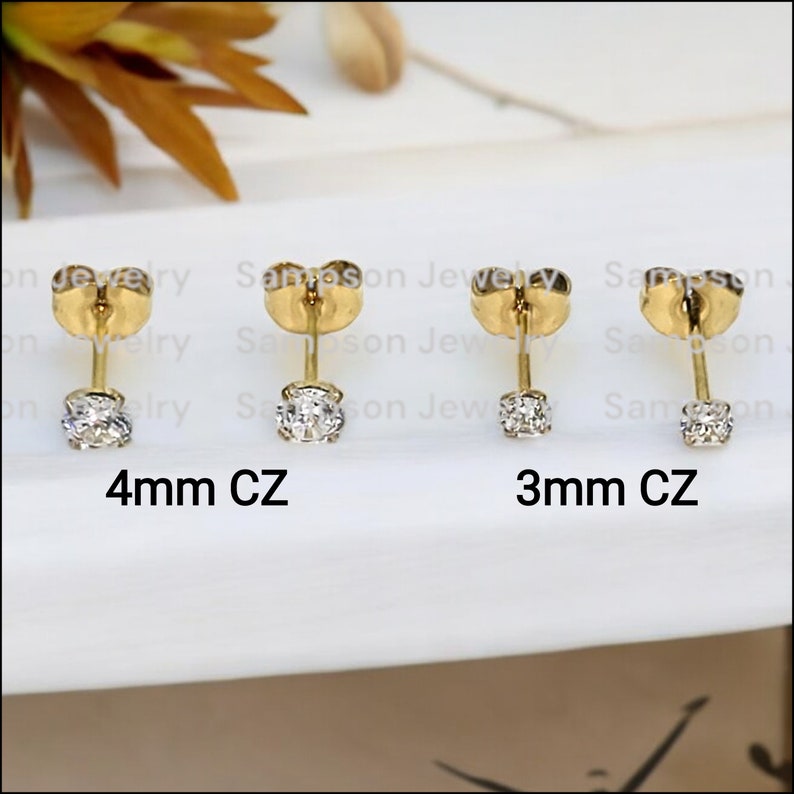 May include: Two pairs of gold stud earrings with clear cubic zirconia stones. The earrings are shown on a white surface. The left pair has 4mm stones and the right pair has 3mm stones. 4mm CZ and 3mm CZ are written below each pair of earrings.