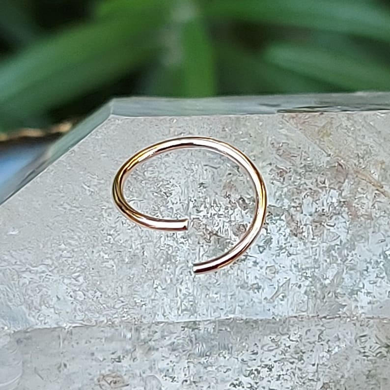 May include: A gold nose ring hoop, a type of body jewelry, is shown on a white crystal surface.