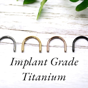 May include: Four D-shaped metal rings in silver, gold, rose gold, and black. The text "Implant Grade Titanium" is printed below the rings. The rings are displayed on a white wooden surface with green foliage in the background.