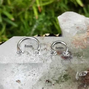 May include: Two silver septum rings with clear cubic zirconia stones. The ring on the left is 8mm in diameter and the ring on the right is 6mm in diameter.