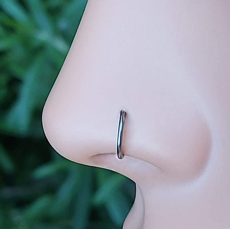 May include: A close-up of a silver nose ring hoop on a mannequin's nose. The ring is simple and understated, with a smooth finish.