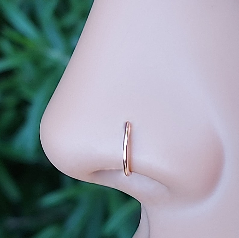 May include: A close-up of a rose gold nose ring hoop. The hoop is thin and delicate, and it is worn in the left nostril.