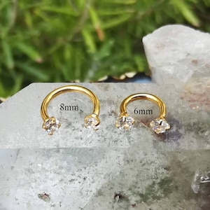 May include: Two gold-toned circular hoop earrings with clear gemstones. The hoop on the left is 8mm in diameter and the hoop on the right is 6mm in diameter.