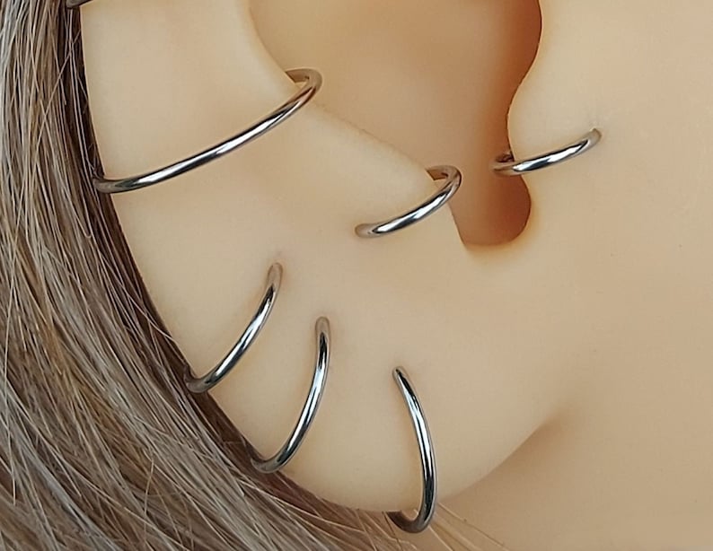 May include: Close-up of a person's ear with multiple silver hoop earrings. The earrings are stacked on top of each other, creating a layered look.