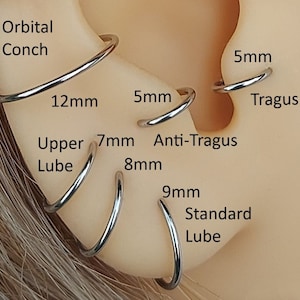 May include: Close-up of an ear with multiple hoop earrings in different sizes. The earrings are labeled with their corresponding sizes in millimeters: 5mm, 12mm, 7mm, 8mm, 9mm. The ear piercings are labeled: Orbital Conch, Tragus, Anti-Tragus, Upper Lube, Standard Lube.