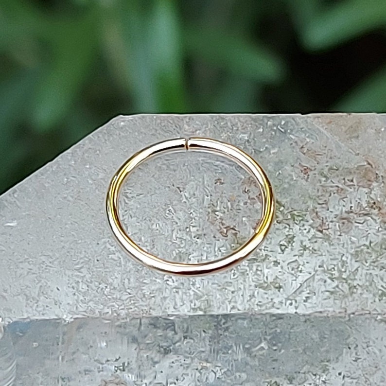 May include: A gold nose ring hoop, a type of body jewelry, is shown on a white crystal surface.