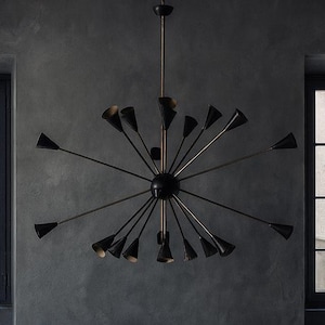 Op de afbeelding: Een modern, donker interieur met een zwarte kroonluchter met meerdere kegelvormige lichten. Daaronder staat een donkere houten kast met lades en een plank op wielen. Twee ramen flankeren de kast en de muren zijn donkergrijs.