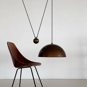 May include: An interior design scene featuring a brown dome pendant light, a brown chair with black legs, and a hanging counterweight. The light fixture and chair are set against a white wall, evoking a mid-century modern style.