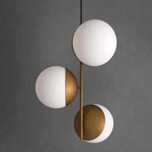May include: A modern pendant light fixture featuring three spherical shades. Two shades are half-gold and half-white, while the third is a full white sphere. The fixture has a gold-colored metal frame and hangs from a black cord.