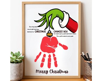 Könnte beinhalten: Ein gerahmter Weihnachtsdruck mit einem roten Handabdruck und einer grünen Grinch-Hand, die einen Weihnachtsbaumschmuck hält. Der Text lautet "Frohe Weihnachten" und enthält festliche Feiertagsphrasen.
