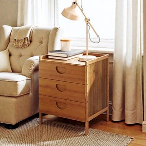 May include: A light brown wooden nightstand with three drawers and a top shelf. A beige lamp, a book, and a small white jar with a wooden lid sit on top. The nightstand is next to a beige armchair.