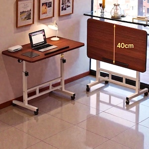 May include: A brown adjustable height desk with a rectangular tabletop. The desk has a white metal frame with wheels and a height of 40cm (15.7 inches). A laptop, tablet, and lamp are on the desk. The desk is next to a window.