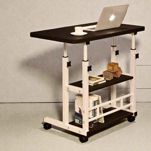 May include: A black and white adjustable laptop desk with a black tabletop and two shelves. A laptop and a cup and saucer are on the top. The desk has wheels for mobility. Books and decorative boxes are on the shelves.