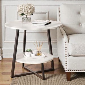 May include: A white marble-topped side table with a lower shelf, supported by dark brown metal legs. The table holds a vase of white flowers, books, and a tray with decorative items. A cream-colored armchair is partially visible.