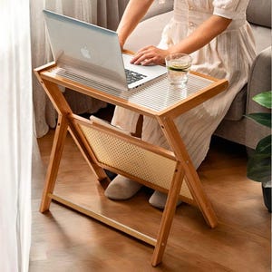 May include: A folding bamboo laptop table with a clear glass top and a woven rattan shelf. The table has an X-frame design. A laptop and a glass of water are on the table. The table is on a wooden floor.