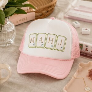 May include: A light pink and white trucker hat featuring a graphic of four mahjong tiles arranged in a row. Each tile displays a single letter, spelling out the word MAHJ in brown font, with small blue bow illustrations at the base of each tile.