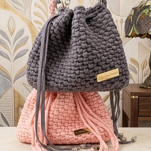 May include: A dark gray crochet bucket bag with a drawstring closure and a gold-toned metal tag reading A Twisted Stitch, stacked atop a light pink crochet bucket bag with matching drawstring cords and silver-toned metal bead accents at the ends.