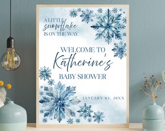 Puede incluir: Un letrero enmarcado para baby shower con un tema invernal. El letrero presenta copos de nieve azules de acuarela y el texto "A little snowflake is on the way" y "Welcome to Katherine's Baby Shower". También se incluye la fecha "02 de enero de 20XX".