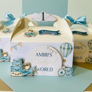 Może przedstawiać: Trzy kremowe pudełka na upominki z niebieskimi akcentami, każde ozdobione niebieską kokardą, chmurą i samolotem. Pudełka zawierają tekst "AMIRI'S Wonderful WORLD" oraz imiona "Santiago" i "Eva".
