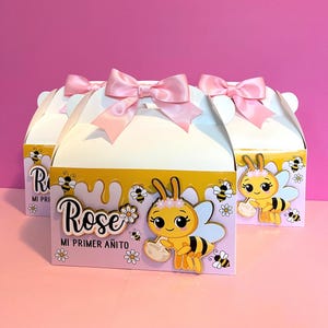 Op de afbeelding: Witte zadeldakdozen met roze strikken, versierd met tekenfilm bijen en de naam "Rose". De dozen hebben een roze en geel ontwerp met de tekst "MI PRIMER AÑITO".