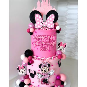 Puede incluir: Una tarta de cumpleaños rosa de dos pisos decorada con elementos temáticos de Minnie Mouse. La tarta presenta un gran lazo con lunares blancos, recortes de Minnie Mouse y el texto "Happy 2nd Birthday Eila". La tarta está adornada con bolas decorativas rosas, negras y blancas.