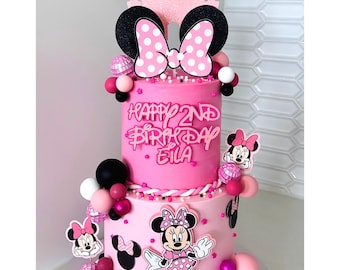 May include: A two-tiered pink birthday cake decorated with Minnie Mouse themed elements. The cake features a large bow with white polka dots, Minnie Mouse cutouts, and the text "Happy 2nd Birthday Eila". The cake is adorned with pink, black, and white decorative spheres.
