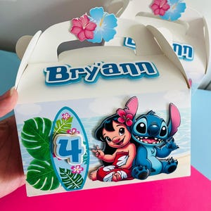 May include: White party favor box with a handle, decorated with a cartoon image of Lilo and Stitch. The box has a blue name, "Bryann", and a surfboard with the number "4". Pink and blue hibiscus flowers adorn the top.