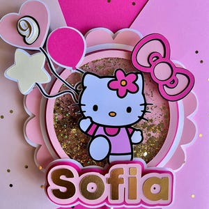 May include: A layered pink and white cake topper featuring a cartoon cat character with a pink flower, balloons, and a heart. The name "Sofia" is displayed in gold lettering. The topper is decorated with gold glitter.