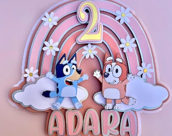 Peut inclure: Décoration de gâteau multicouche avec un arc-en-ciel, des marguerites et deux chiens de dessin animé. Le chiffre "2" est en jaune et le nom "ADARA" est en rose. Le fond est rose clair.