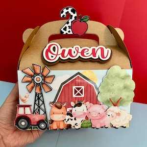 Può includere: Una scatola regalo di cartone marrone a tema fattoria. La scatola presenta un manico e il nome "Owen" in rosso e bianco. La scatola è decorata con un fienile rosso, animali della fattoria, un mulino a vento e un trattore. Un numero "2" e una mela sono in cima.