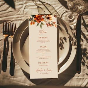 May include: A wedding menu card with floral accents in orange, white, and green. The menu lists entree, main, and dessert options. The card is on a white plate with gold cutlery. The menu is for Amelia & James, October 25, 2022.