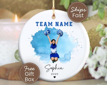 May include: A round white ceramic ornament featuring a blue watercolor splash design. In the center, a cheerleader in a blue and white uniform jumps with pom-poms. The text reads TEAM NAME, Sophia, and 2025 with a small heart icon below.