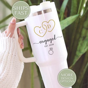 May include: White insulated tumbler with a handle, featuring gold heart-shaped initials and the word "engaged" with "EST. 2026" and a ring graphic. The tumbler is held against a background of green foliage.
