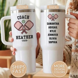 May include: A white stainless steel tumbler with a handle and straw. The front features two pink pickleball paddles, a green ball, and the text COACH HEATHER -2026-. The back displays a small pickleball graphic and a list of names: SAMANTHA, ABIGAIL, KYLIE, JENNIFER, OLIVIA, SOPHIA.