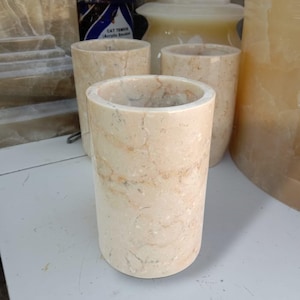 May include: Three cylindrical containers crafted from polished beige marble with natural veining and cream-coloured swirls. Each vessel features a smooth, straight-walled exterior and a hollow interior, showcasing the unique stone patterns and textures.