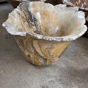 May include: A large, hand-carved stone bowl featuring natural swirling patterns in shades of cream, tan, brown, and grey. The bowl has a polished interior and a rough, organic, uneven rim that highlights the natural texture of the stone material.