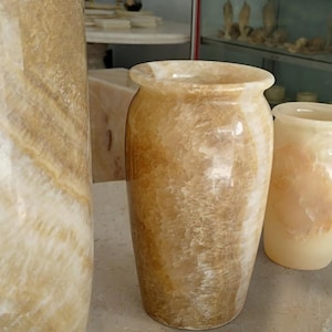 May include: A cylindrical vase crafted from polished honey-colored onyx stone, featuring natural translucent bands and swirls of cream, tan, and golden brown. The vessel has a smooth, glossy finish with a wide, rounded rim and a solid, stable base.