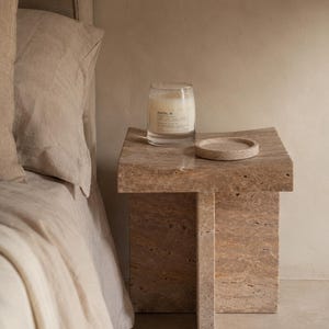 May include: A beige travertine stone side table with a cross-shaped base and a flat square top. On the table sits a clear glass candle jar with a white label reading SANTAL 26 and a small circular stone coaster next to the candle.