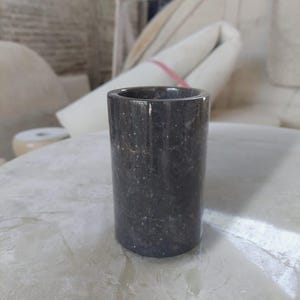 Può includere: Un contenitore cilindrico realizzato in pietra grigio scuro con una finitura lucida e sottili macchioline bianche su tutta la superficie. L'oggetto presenta un design liscio a pareti dritte con interno cavo e base circolare piatta.