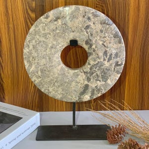 May include: A circular, gray stone sculpture with a central hole, mounted on a black metal stand. The stone has a mottled pattern of gray and beige. The sculpture is a decorative item.
